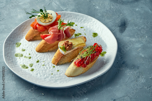 Assorted bruschetta with baked peppers, camembert, prosciutto and lightly salted salmon in a white plate on a gray background