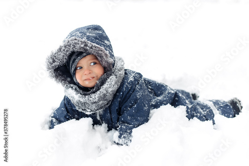 Close-up image of adorable baby boy in a warm white snow suit playing and lying in snow with cute face.
