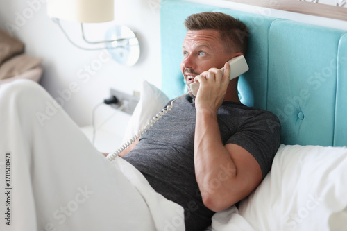 Man lies in bed in hotel room and talk on phone. Calling hotel staff and customer service remotely.