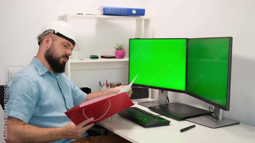 Engineer Wearing Safety helmet or Hardhat Works on a Computer, Two Monitor Screens Show Green Screen Displays. remote work at home Engineer Wearing Safety helmet or Hardhat Works on a Computer, Two Monitor Screens Show Green Screen Displays. remote work at home