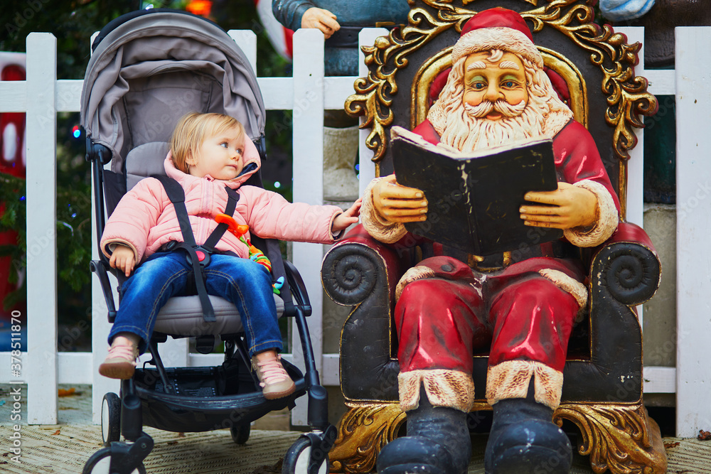 Adorable toddler girl sitting in pushchair near wooden Santa Claus