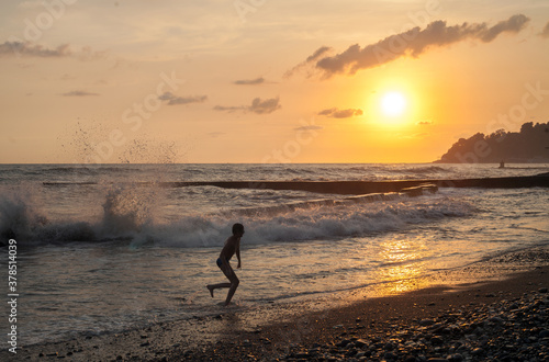 A child running along the seashore against the backdrop of storm waves and the setting sun.