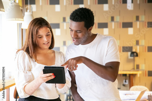 African American man pointing at tablet in colleagues hands. Brunette young woman holding device, standing indoors, showing something, smiling and talking. Communication, design and teamwork concept