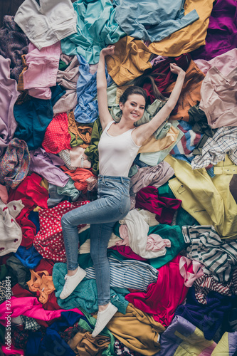 High angle above view vertical photo of cheerful funny lady spring cleaning household lying many clothes heap stack floor shopper surrounded sales stuff do not care mess spread hands indoors