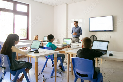 Confident teacher explaining lesson to pupils. Multiethnic children sitting at table in classroom, listening middle-aged man and using laptop computers. Childhood and digital education concept