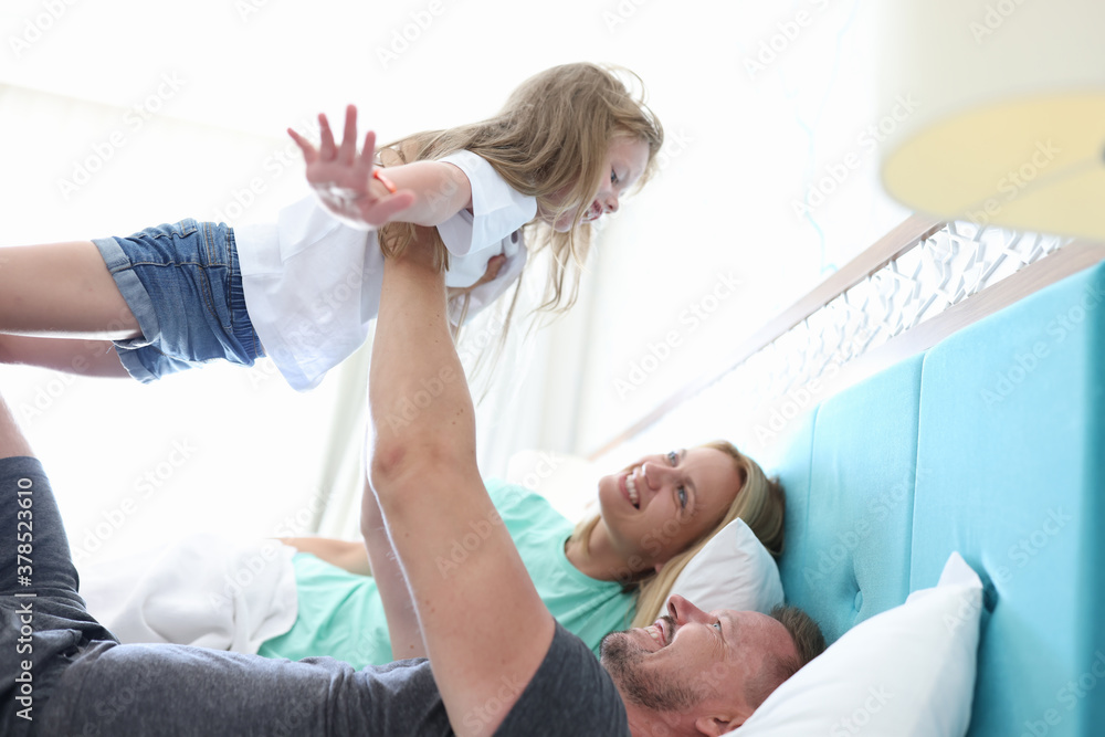 Fototapeta premium Man and woman lie side by side in bed and look up at their daughter. Dad hold girl outstretched arms up. Child rest directly on man arms with his arms out to sides.