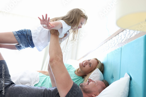 Man and woman lie side by side in bed and look up at their daughter. Dad hold girl outstretched arms up. Child rest directly on man arms with his arms out to sides.