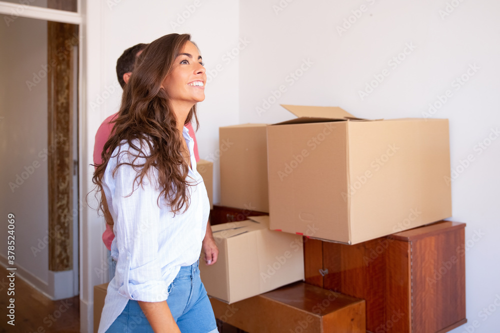 Cheerful excited woman and her boyfriend looking over their new apartment with carton boxes and furniture. Copy space. New home concept Cheerful excited woman and her boyfriend looking over their new apartment with carton boxes and furniture. Copy space. New home concept