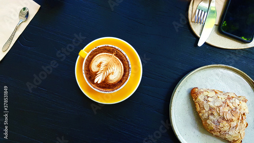 Top view of art latte coffee with almond cream croissant, knife, fork and phone on blue wooden background with copy space at cafe. Flat lay of hot cappuccino drinking. Relaxing time in the morning