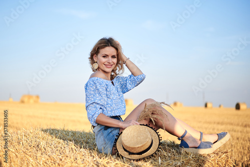 Schilderij op canvas Young curly blond woman, wearing jeans shorts and light blue shirt, holding straw hat, sitting on bale on field in summer