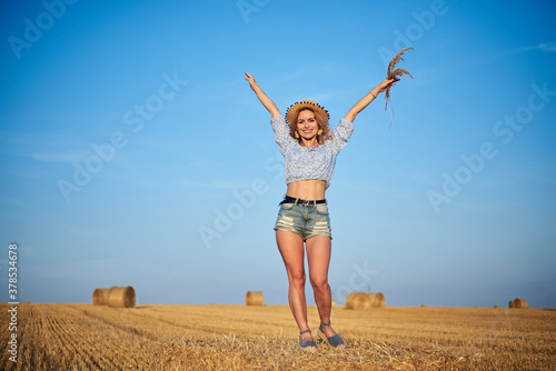 Young curly blond woman, wearing jeans shorts and light blue shirt, holding straw hat, standing on bale on field in summer. Female portrait in natural rural scene. Environmental eco tourism concept.