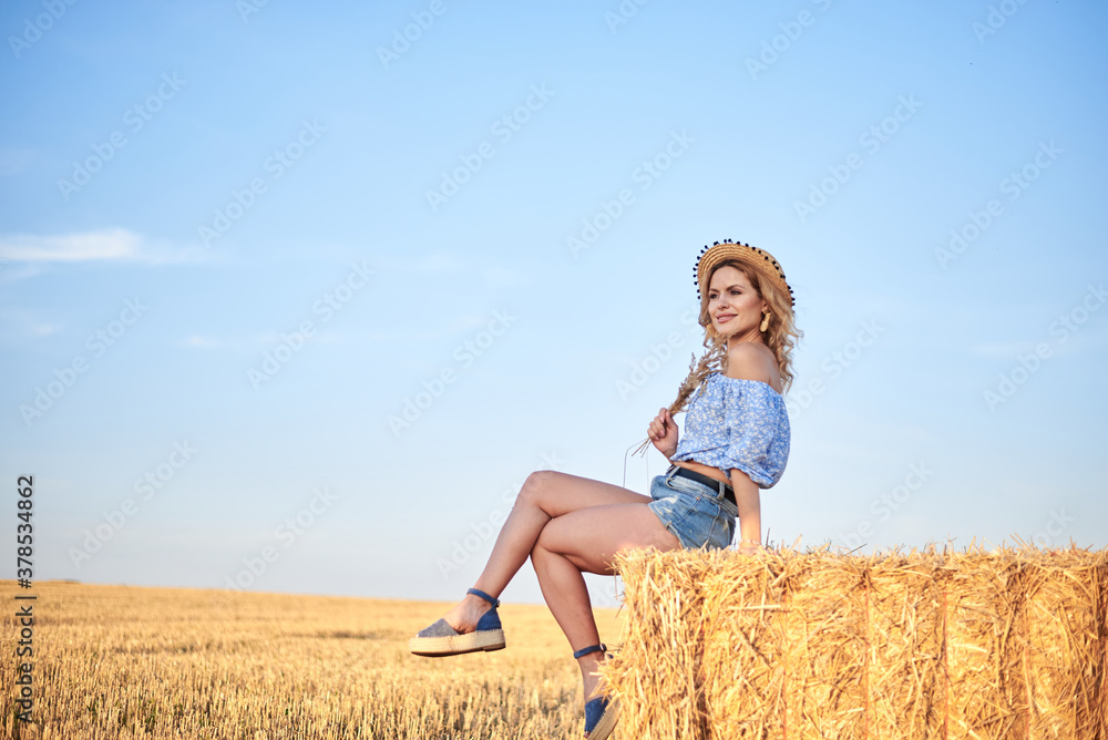 Young curly blond woman, wearing jeans shorts, light blue shirt and straw hat, sitting on bale on field in summer. Female portrait in natural rural scene. Environmental eco tourism concept. Young curly blond woman, wearing jeans shorts, light blue shirt and straw hat, sitting on bale on field in summer. Female portrait in natural rural scene. Environmental eco tourism concept.