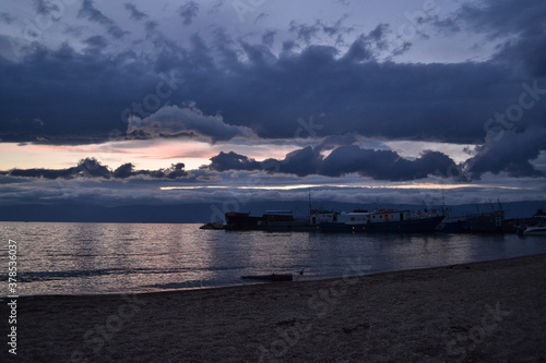 light pink sunset with blue dark clouds in sky in bay of lake Baikal with jetty mole pier with old ships and boats, evening sea