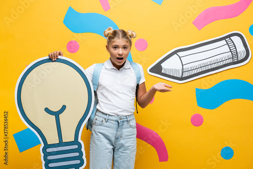 Confused schoolgirl pointing with hand while holding paper light bulb bear paper art on yellow background