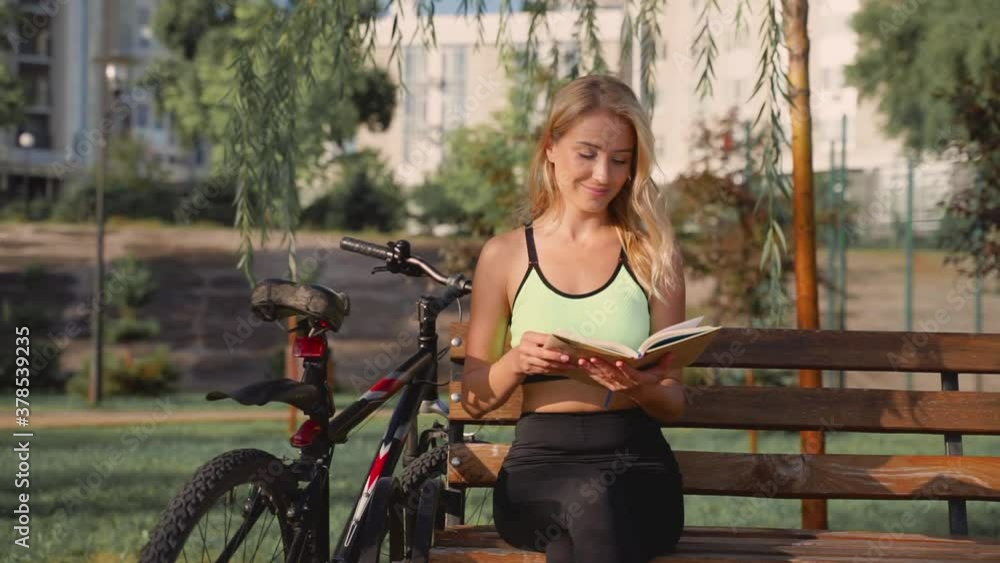Happy woman reading a book, she relaxes and enjoys free time on a sunny afternoon. Female athlete sitting on a bench next to her bike outdoor. Happy woman reading a book, she relaxes and enjoys free time on a sunny afternoon. Female athlete sitting on a bench next to her bike outdoor.
