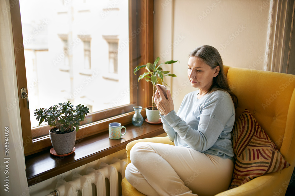 Casually dressed serious retired female sitting comfortably in yellow armchair by window using wireless internet connection on smart phone, reading or texting message, browsing social networks Casually dressed serious retired female sitting comfortably in yellow armchair by window using wireless internet connection on smart phone, reading or texting message, browsing social networks