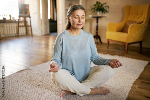 Calm barefoot middle aged female sitting on carpet in half lotus posture, making mudra gesture and closing eyes, having peaceful facial expression, doing mindful meditation, concentrating on breathing