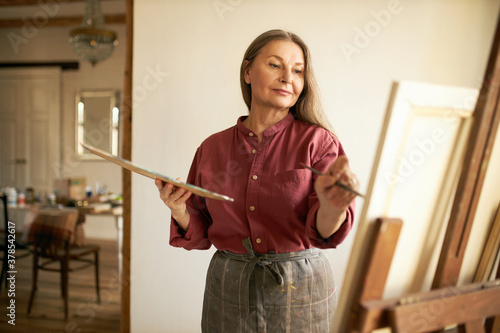 Mature people, aging, hobby and leisure. Indoor image of good looking concentrated senior woman in apron using palette to mix oil pigments, applying paint on canvas using brush, making painting