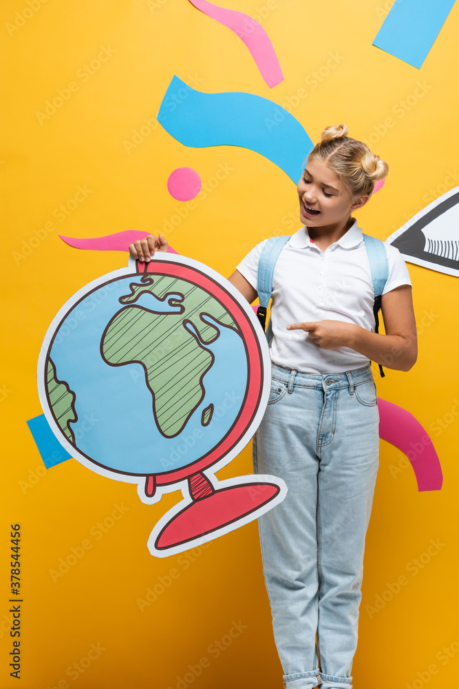Fototapeta premium excited schoolgirl pointing with finger at globe maquette near paper cut pencil and colorful elements on yellow