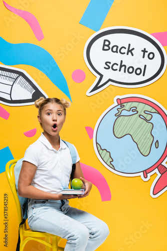 Shocked schoolkid holding book and apple on chair near paper art and speech bubble with back to school lettering on yellow background