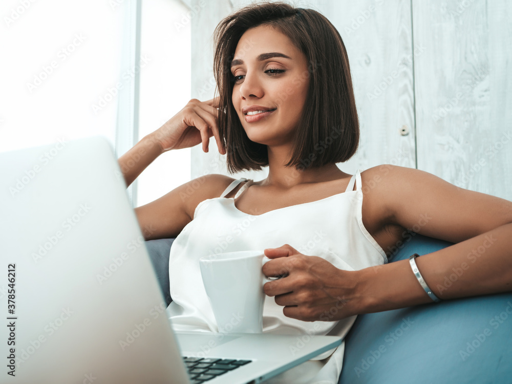 Portrait of beautiful smiling woman dressed in white pajamas. Carefree model sitting on soft bag chair and using laptop. Sexy female reading news and enjoying her morning at balcony, drinking coffee Portrait of beautiful smiling woman dressed in white pajamas. Carefree model sitting on soft bag chair and using laptop. Sexy female reading news and enjoying her morning at balcony, drinking coffee