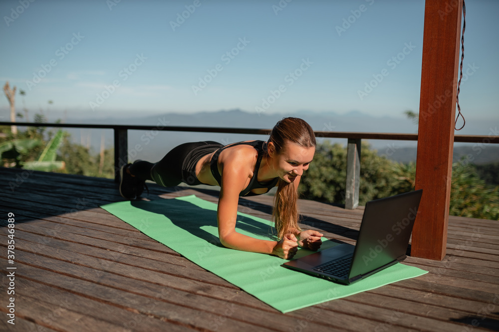 Girl training at home, doing plank and watching videos on laptop, training on terrace with mountain nature view