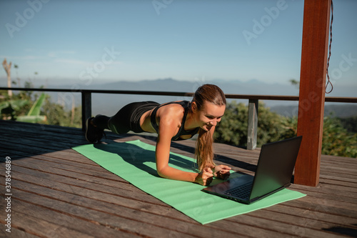Girl training at home, doing plank and watching videos on laptop, training on terrace with mountain nature view photo