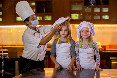 Kid cooking class. Chef putting hats on sudent heads. Two little girls and teacher chef in kitchen during master class learning how to make pizza photo