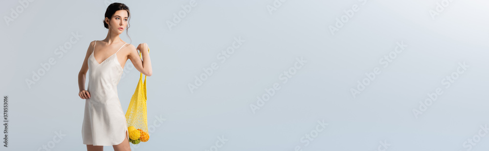 horizontal image of brunette woman in silk dress holding string bag with fruits on grey, ecology concept horizontal image of brunette woman in silk dress holding string bag with fruits on grey, ecology concept