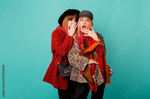 Two stylish blond women posing in studio on blue turquoise background. Friends gossiping and having fun together. Wearing leopard print faux fur coat and wool scarf. Winter fashion.