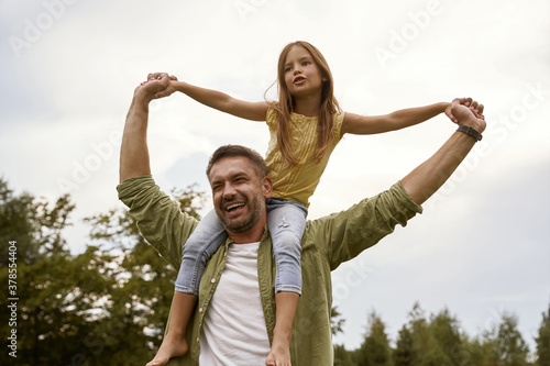 Enjoying time together. Little excited girl having fun with father in park, sitting on his shoulders and smiling