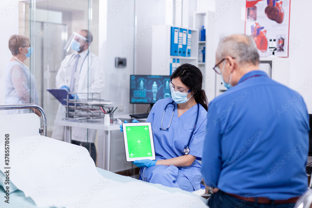 Patient looking at tablet with green screen during consultation in hospital room with nurse wearing face mask against coronavirus as safety precaution. Medical examination for infections, disease . Patient looking at tablet with green screen during consultation in hospital room with nurse wearing face mask against coronavirus as safety precaution. Medical examination for infections, disease .