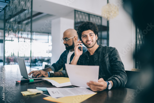 Portrait of cheerful Middle Eastern employer with documents report in hand calling to business partner for discussing strategy while blurred colleague using laptop device for wireless booking