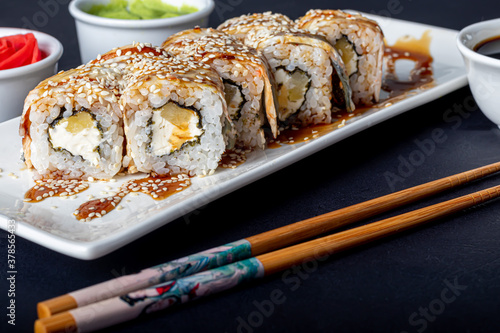 Sushi Sets Uramaki, California, Philadelphia, on a white plate. Nearby ginger and wasabi. Soy sauce in a white bowl. On a dark background.