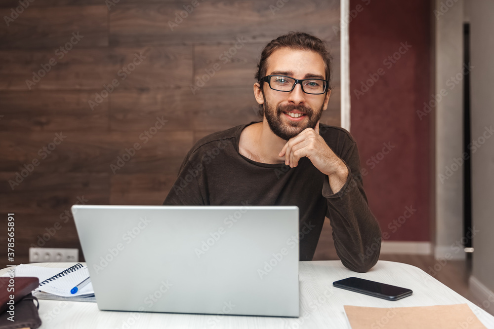 Smiling young man with beard looking at camera while working on laptop Smiling young man with beard looking at camera while working on laptop