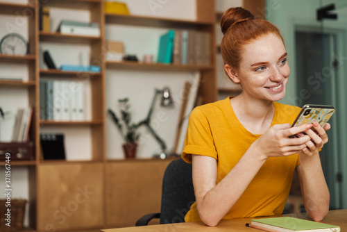 Image of happy redhead girl using cellphone while sitting at table