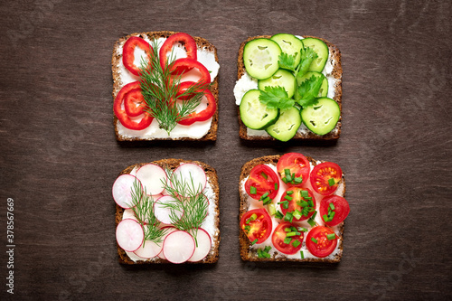 Variety of sandwiches for breakfast - slice of whole grain dark bread, red pepper, cream cheese, cucumbers, radishes, cherry tomatoes, garnished with dill, green onions on dark table Top view Flat lay