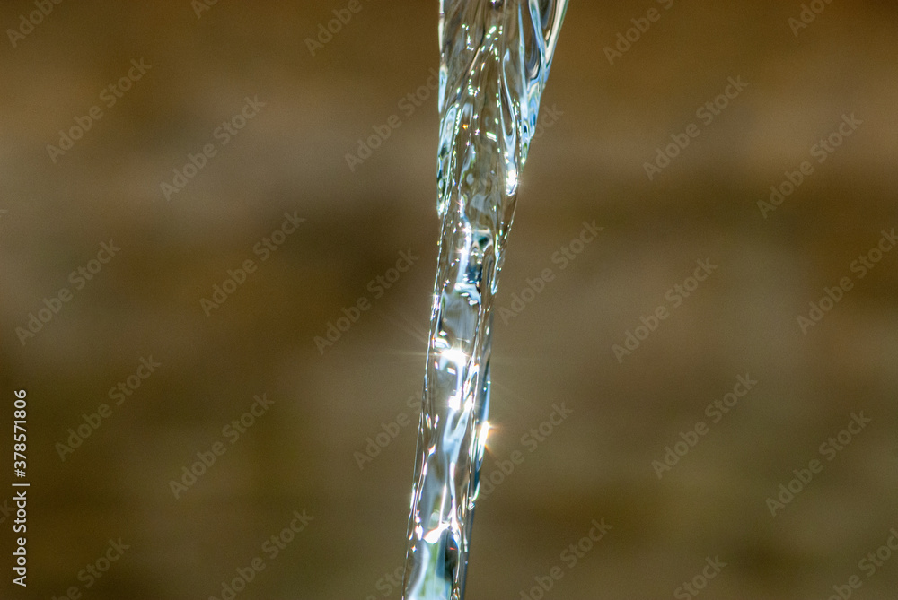 Jet of blue clear water similar to liquid glass shining on a sun light on a blurry brown background