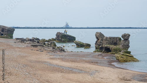Two fishermen standing on the ruins of northern fort in Liepaja, Latvia, and fishing. Abandoned military ruins in Baltic sea on calm summer day. Northern Breakwater and transport vessel in background