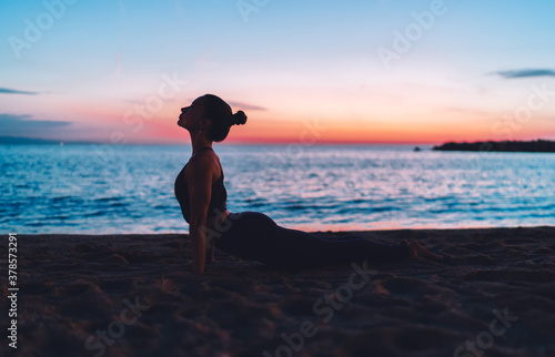 Flexible female yogi practicing cobra pose during evening workout at coastline near ocean horizon enjoying healthy lifestyle  woman with perfect body shape stretching muscles during aerobic pilates