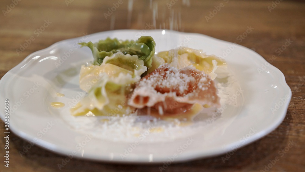 Tortellini on white plate on wooden table, pouring parmesan. Close up tortellini of different colors, green, white, red served with spoon on white plate with melting butter Tortellini on white plate on wooden table, pouring parmesan. Close up tortellini of different colors, green, white, red served with spoon on white plate with melting butter
