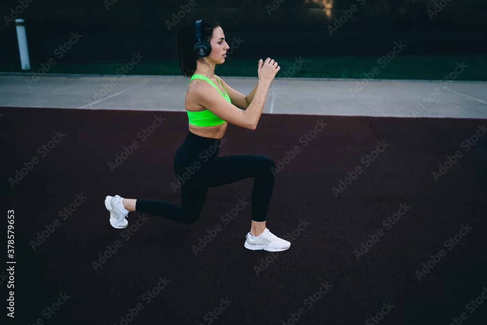 Slim Caucasian girl in bluetooth headphones keeping perfect body shape training legs enjoying online radio via equipment, sportive woman in activewear squatting on stadium during ABT workout Slim Caucasian girl in bluetooth headphones keeping perfect body shape training legs enjoying online radio via equipment, sportive woman in activewear squatting on stadium during ABT workout