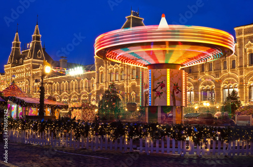 New Year's Fair in Moscow on Red Square. In the foreground is a spinning carousel illuminated by lights. In the background GUM shopping center, decorated with golden lights. Evening.
