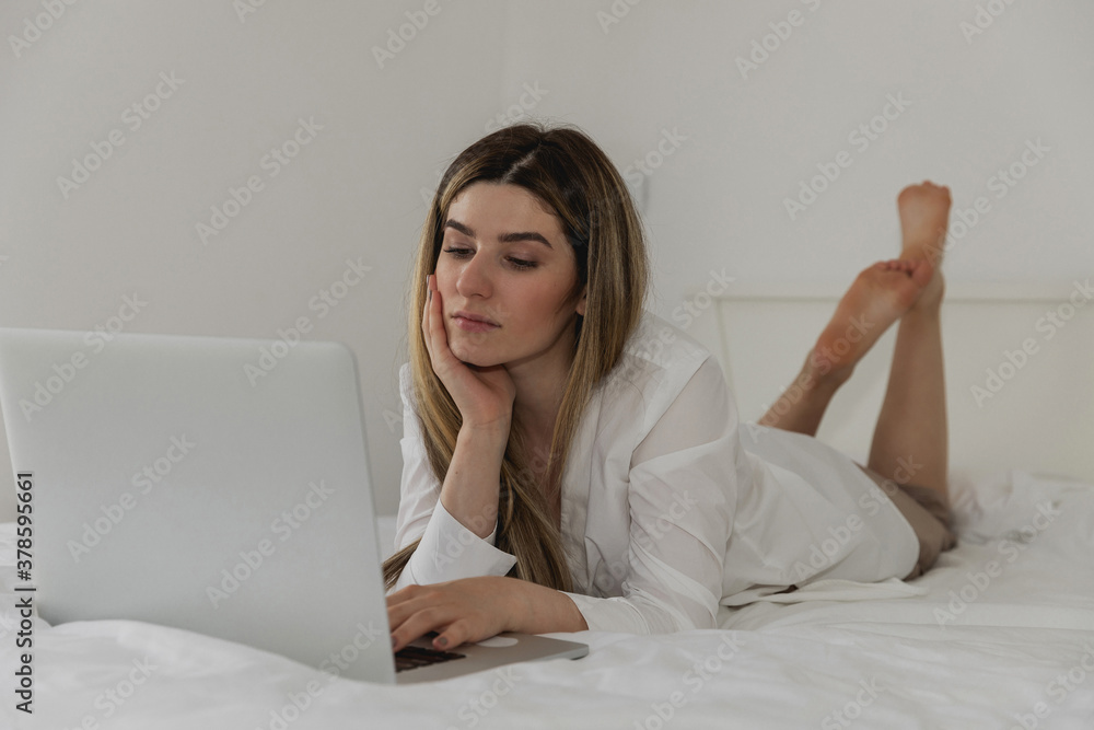 Young beautiful woman relaxing and lies on the bed using laptop computer in the bedroom. Online chat. Working from home. Woman checking social apps. Online order food. Quarantine time. Freelancer Young beautiful woman relaxing and lies on the bed using laptop computer in the bedroom. Online chat. Working from home. Woman checking social apps. Online order food. Quarantine time. Freelancer