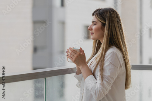 Young woman thinking and looking away with cup of tea or coffee with blurred city background. Girl dressed in white shirt with tea at balcony. Breathing fresh in the morning. Relax comfort concept