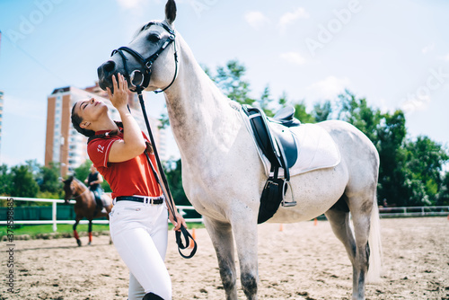 Side view of Caucasian female jockey and white breed horse enjoying friendship during summer training in paddock, youthful girl trying to kiss stallion after professional dressage standing outdoors