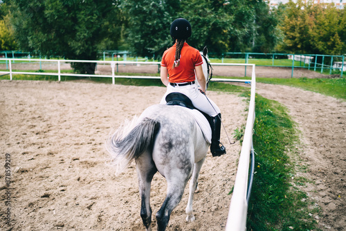 Back view of female horse rider on equestrian sport competition training hobby during recreation leisure, woman in dressed clothing for horseback riding sitting on powerful white stallion