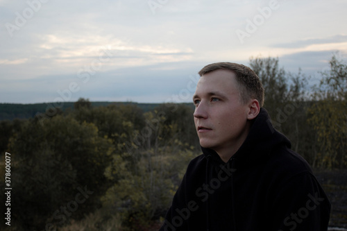 Portrait of a light guy in a black hoodie against the sky and forest. Blonde with blue eyes on top.