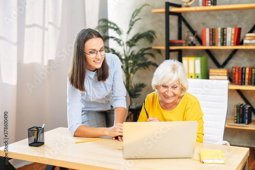 Young manager boss checking work of older female trainee, looking at computer screen in office. Youthful mentor is teaching senior female intern