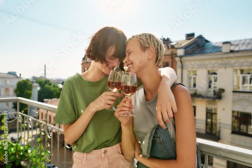 Sticker Two cheerful girls holding glass of wine, hugging while having party, celebrating anniversary on the balcony